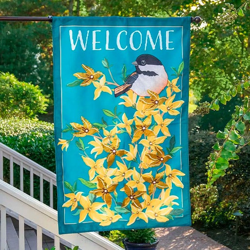 A large estate house welcome flag with chickadee and forsythia, hanging from a front porch