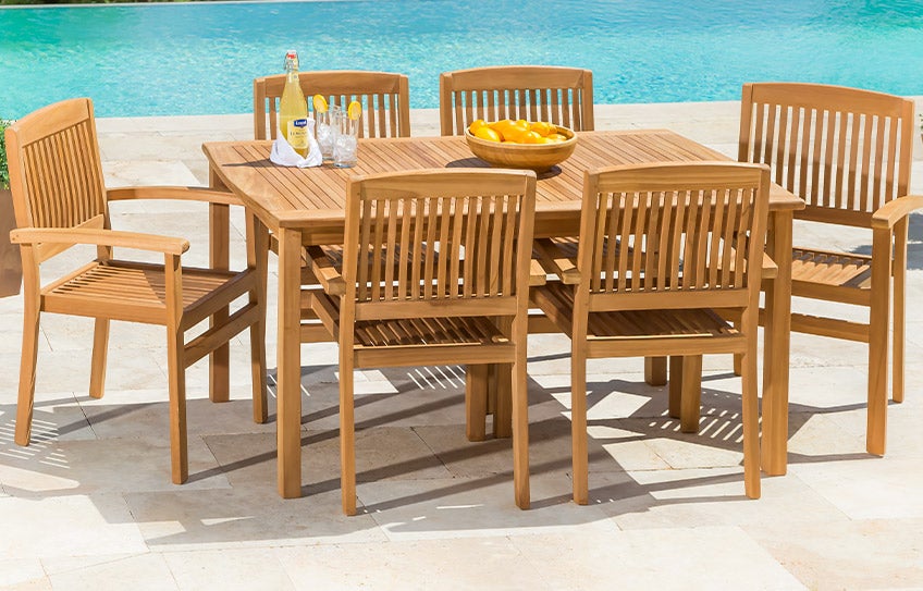 A Teak Collection dining table and six chairs on a poolside patio in the sun.